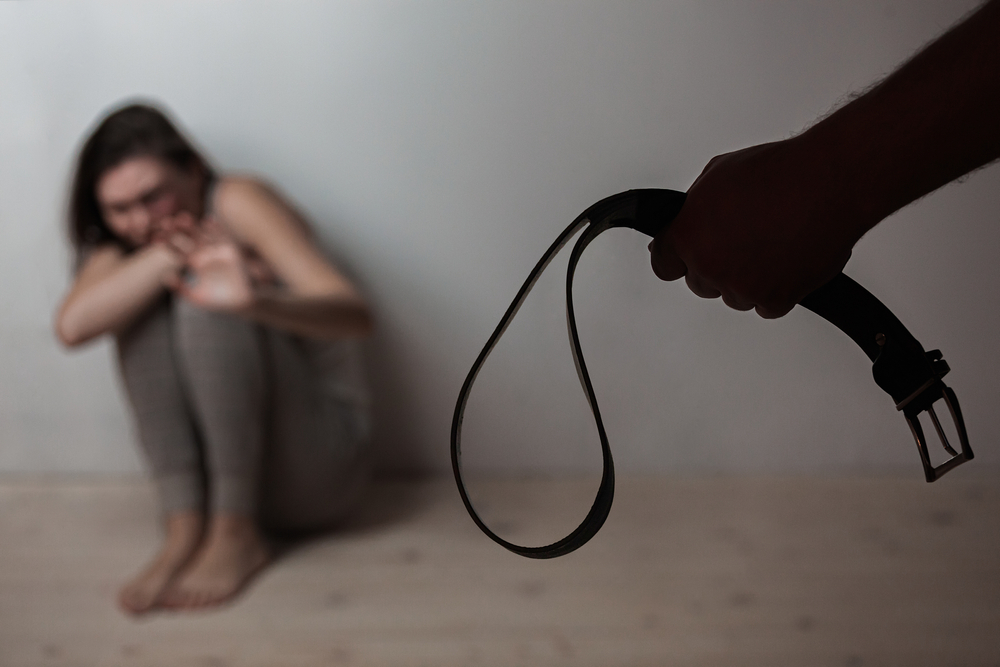 a man holding a belt up to a woman cowering in fear